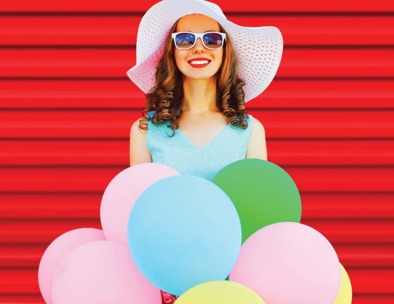 Girl wearing rounded sunglasses with a white hat and a light blue dress holding a bunch of balloons.